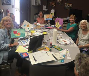 Six women sit around a table scattered with art supplies. Four of the women hold up paintings.