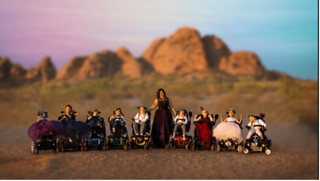 Nine boys and girls in power wheelchairs and colorful outfits pose with Melanie Thompson.